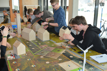 Jugendliche sitzen um einen Tisch und hantieren an kleinen Holzhäuschen herum. Auf der Tischplatte wird ein Miniatur-Dorf aufgebaut. 